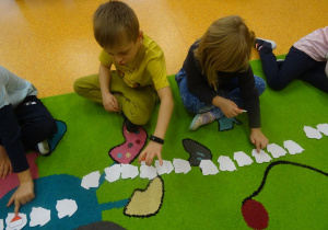 Dwójka dzieci siedzi na dywanie z ułożonymi w szeregu białymi szablonami mapy Polski, przeliczają je.